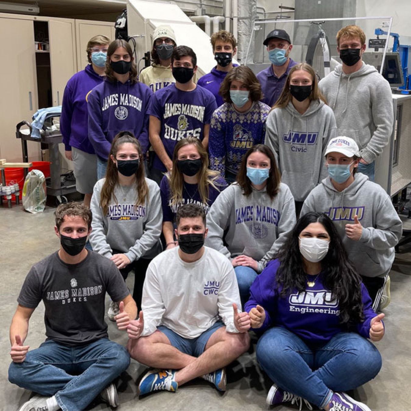 Sixteen students wearing medical masks in a research laboratory. Some are seated; others stand behind them. Most of the people are wearing shirts with the James Madison University logo.