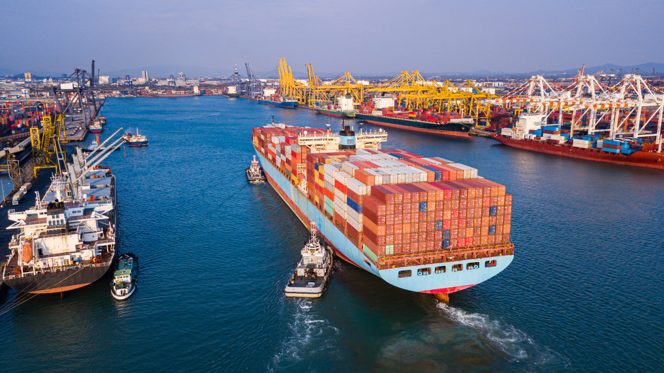 A giant container ship loaded with many goods is brought into a large port by tug boats.