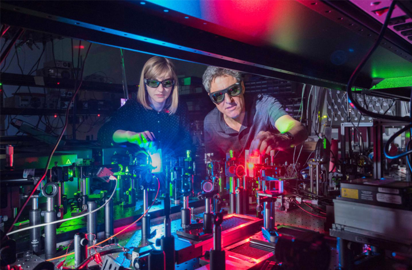 In a dark research laboratory, two scientists wearing goggles lean over a table to adjust lenses reflecting blue and red light from lasers.