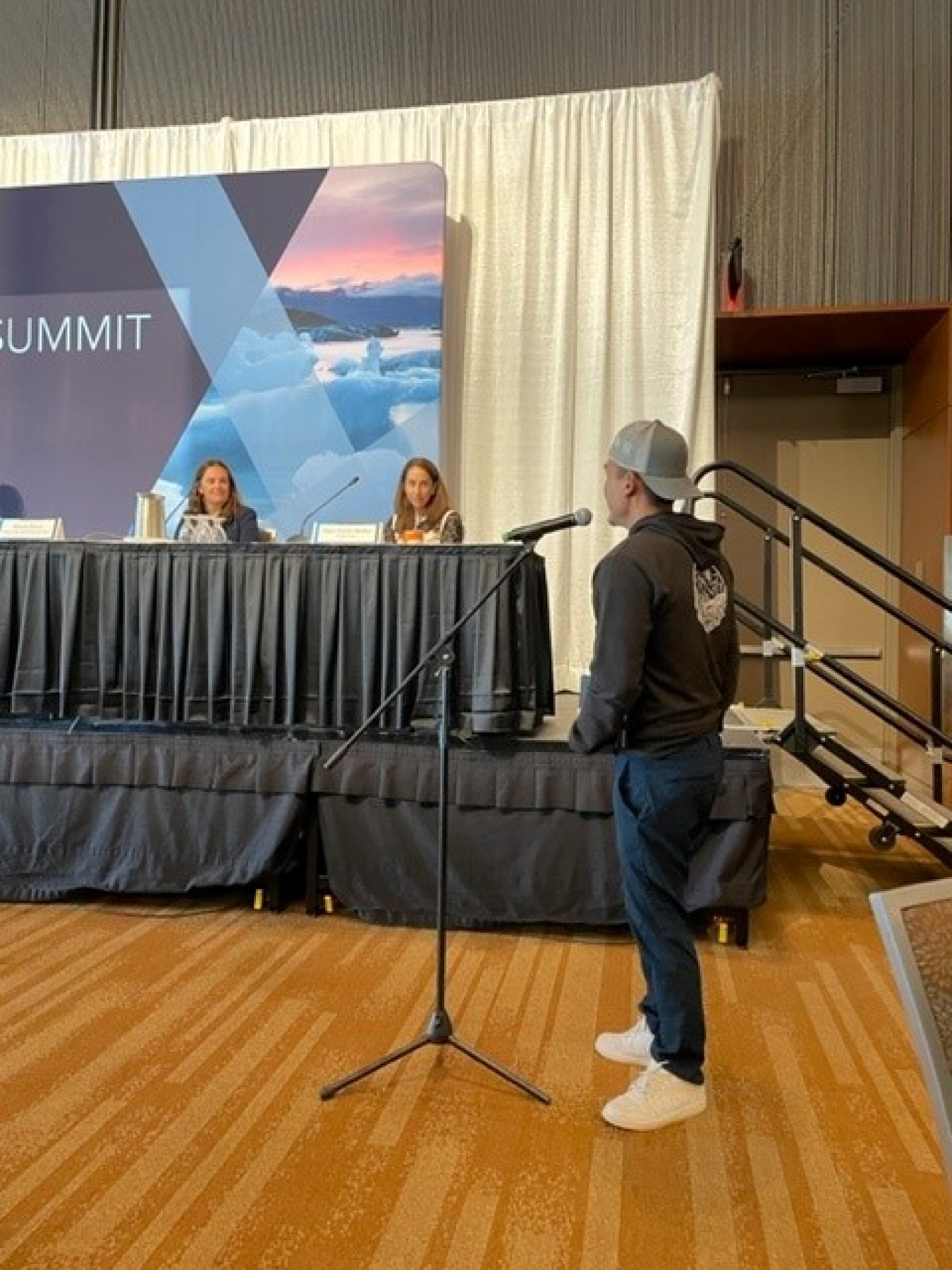 A young man stands at the microphone looking at the panelists at ArcticX. 