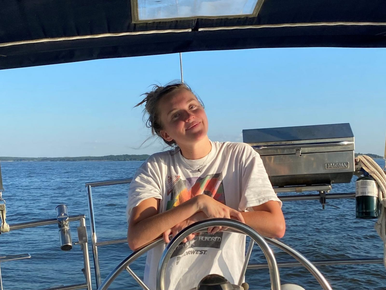 Young woman stands at the wheel of a boat with her arms crossed, surrounded by blue ocean water. She is wearing a white t-shirt, her windblown hair is in a bun, and she is grinning at  the camera with her head tilted slightly.