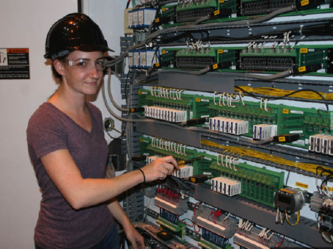 Young woman wearing a hard hat, safety glasses, and a t-shirt grins at the camera while working on machinery.