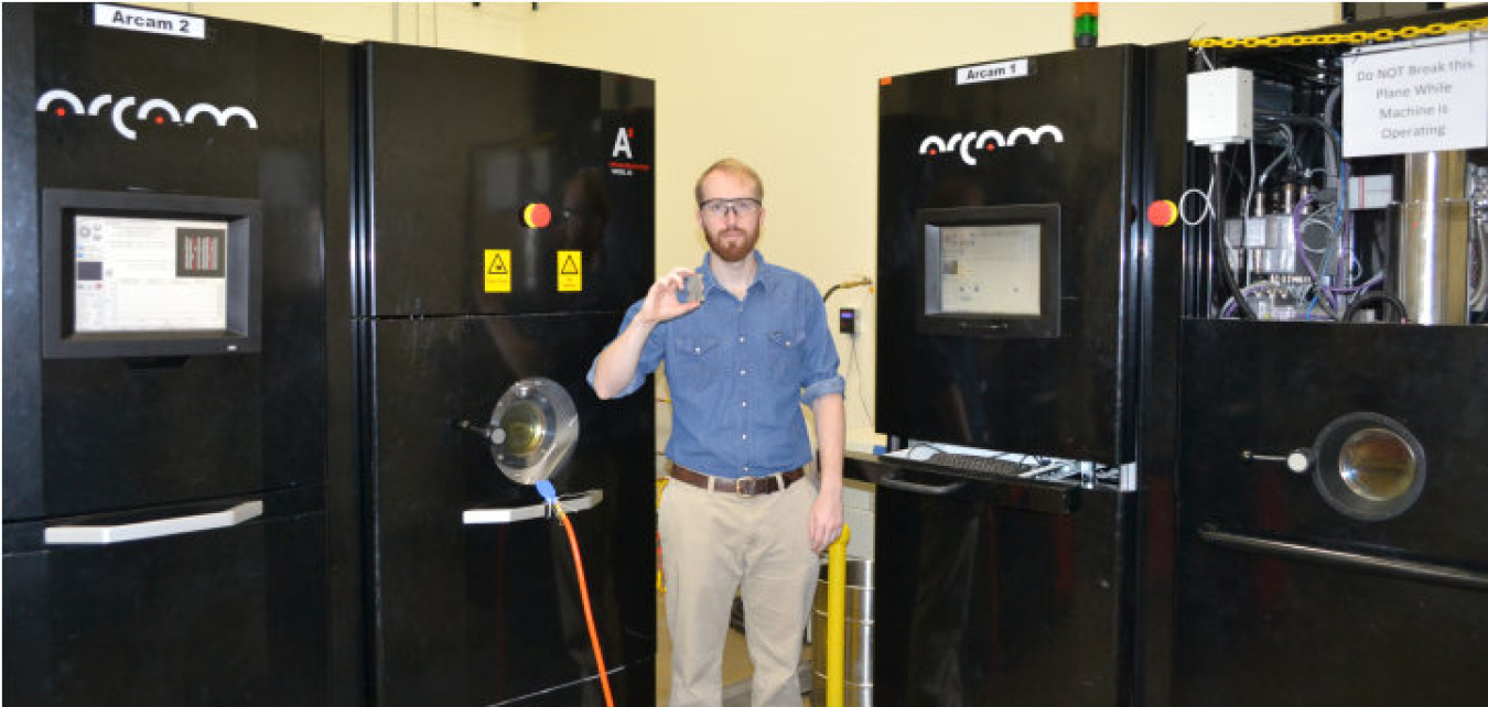 Young man in denim shirt, khaki pants, and safety goggles stands between four large, black machines. He holds a small, rectangular device in one hand at shoulder level.