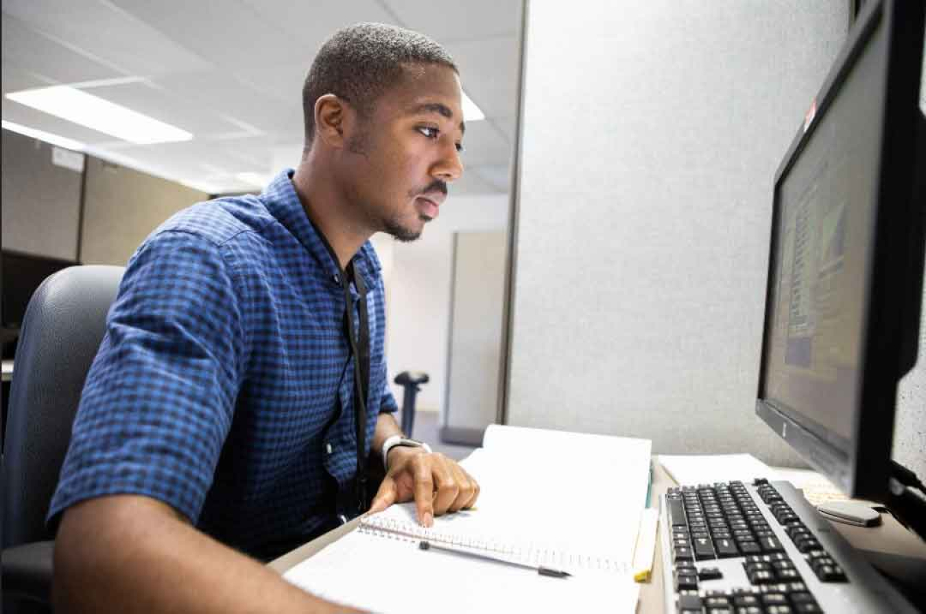 Young man looks in a blue checked shirt sits at a desk looking at computer monitor while his hand rests on notebook.