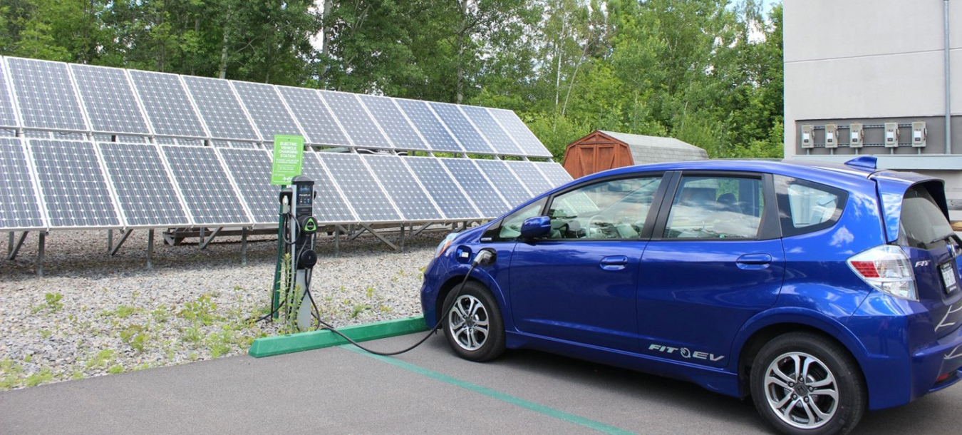 A car plugged into an electric vehicle charge port with a photovoltaic panel behind it.