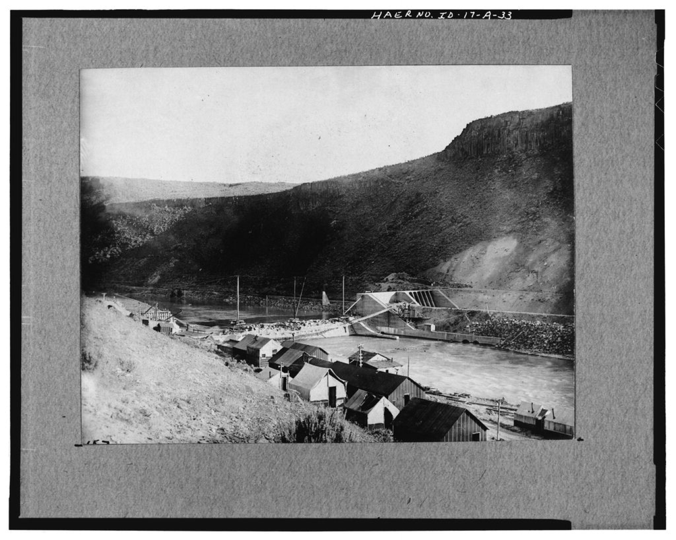 An old black-and-white photo of a dam across a river and small huts on the shore.