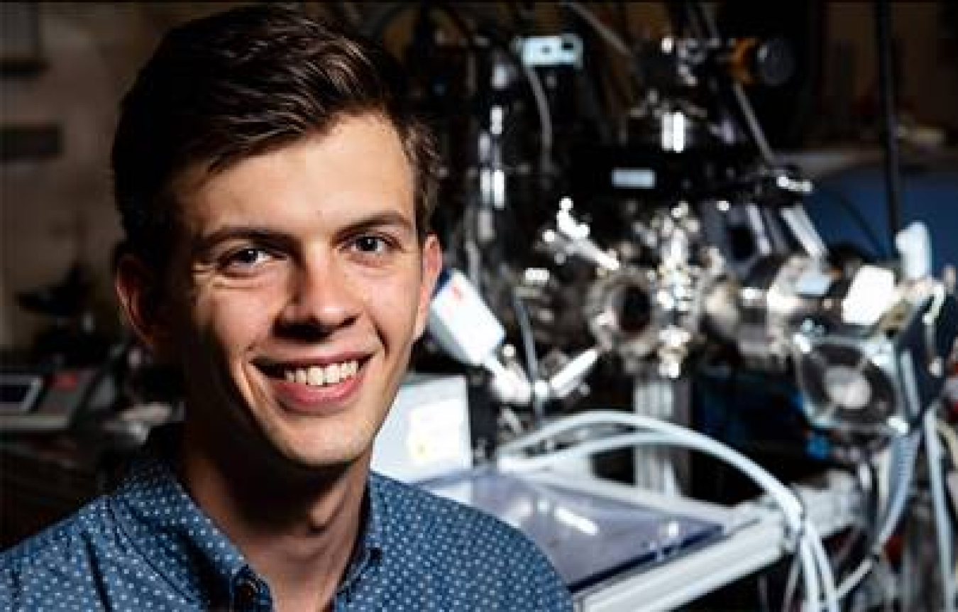 Young man smiles kindly at the camera. He is wearing a blue polka dot button up shirt. Behind him is lab machinery.