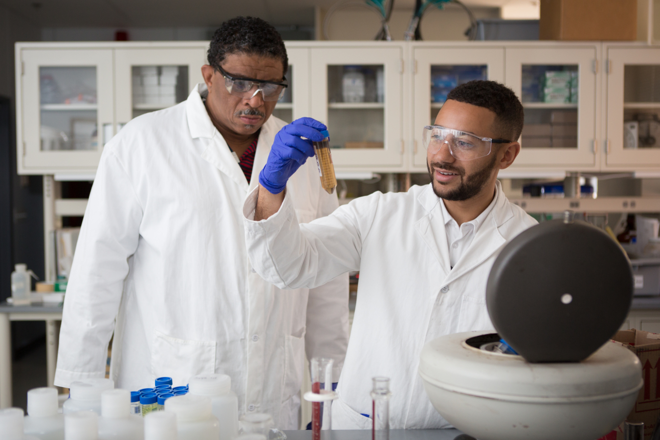 Two scientists, a student and his mentor, wear lab coats and goggles while looking at a conical tube that they've removed from a table top centrifuge