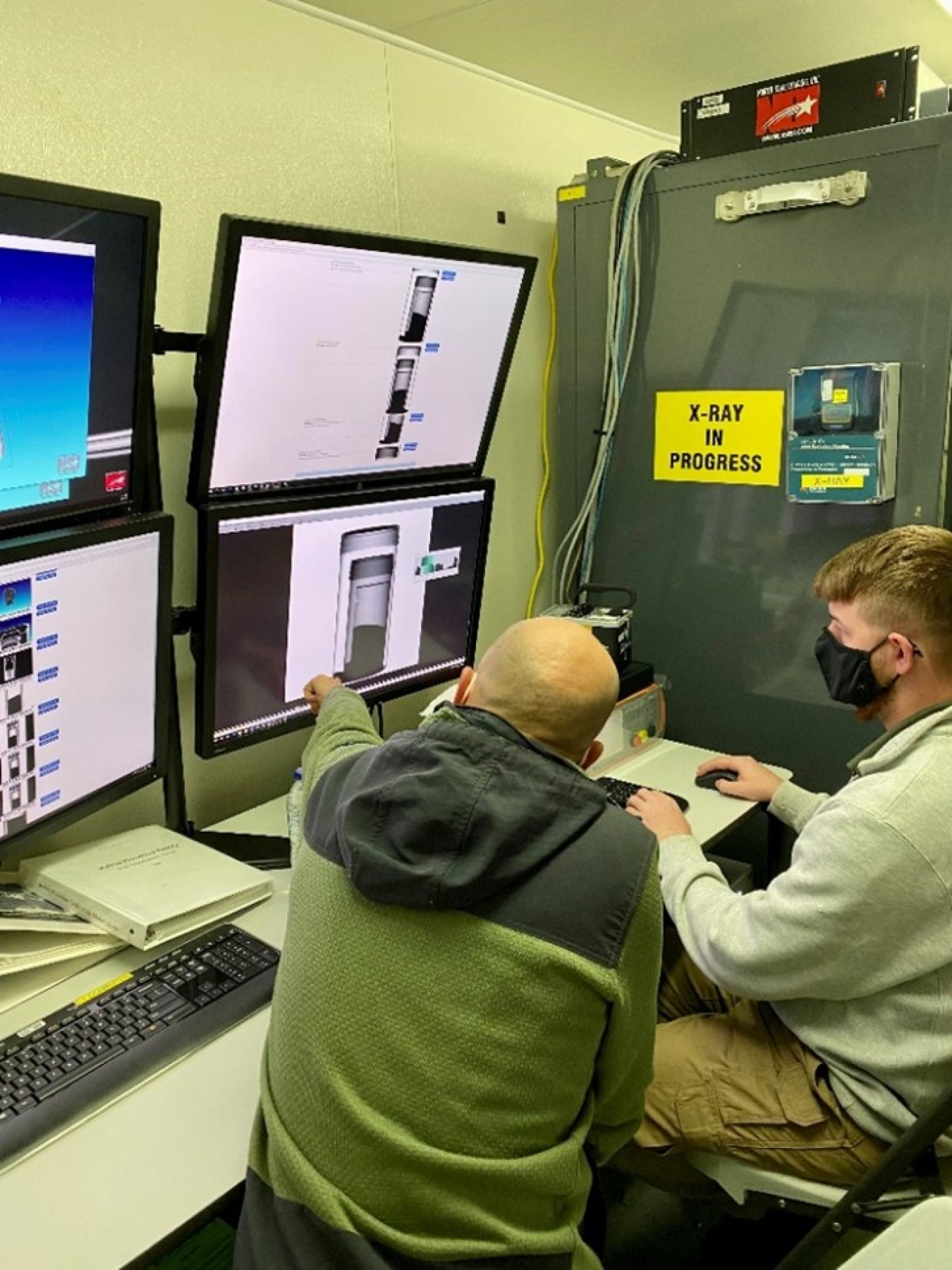 In a photo taken from behind, two men look at computer screens. One man points to an image of canned plutonium on the screen.