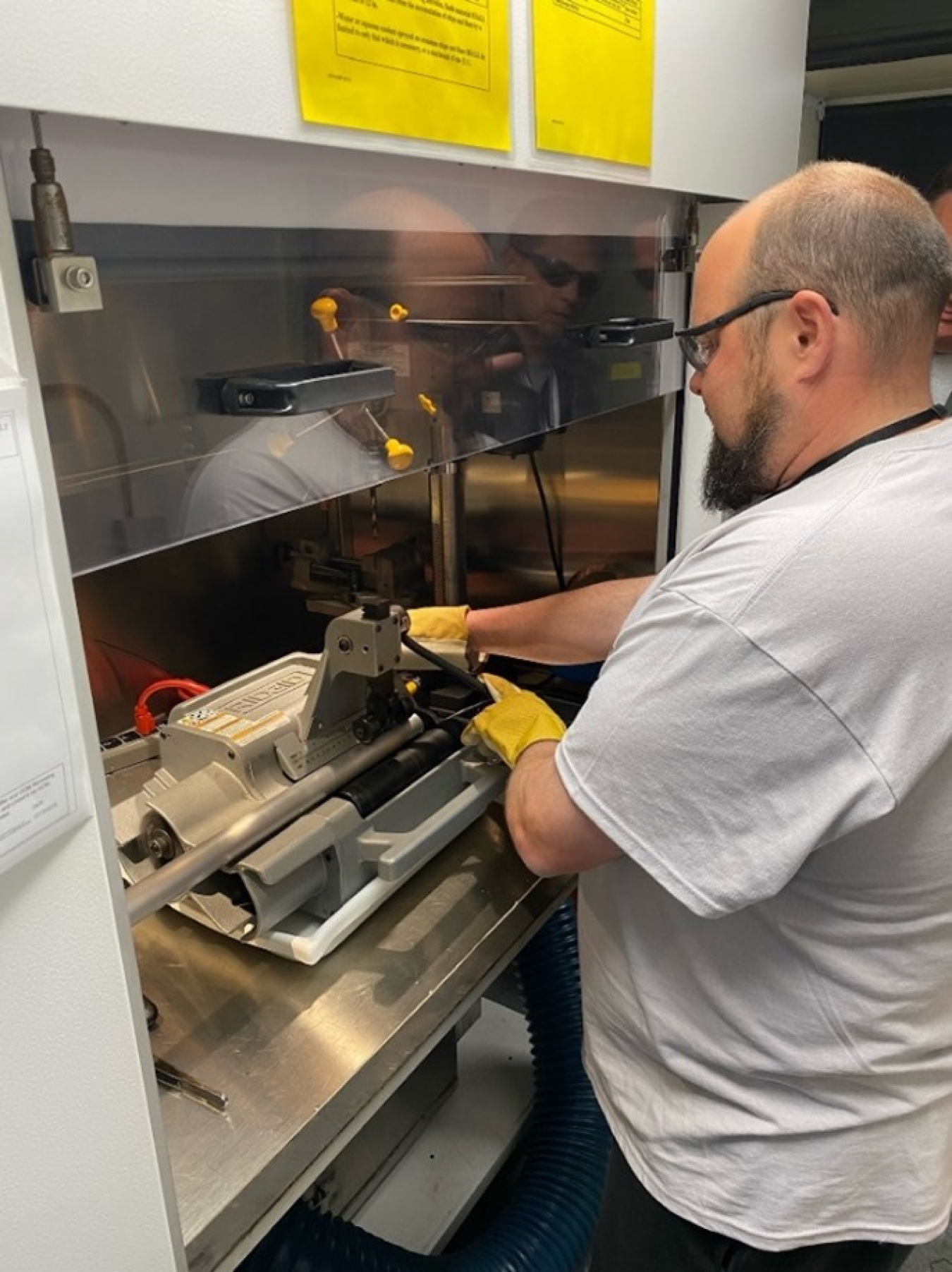 Working in in front of a chemical exhaust hood, a man in rubber gloves operates a machine inside it.