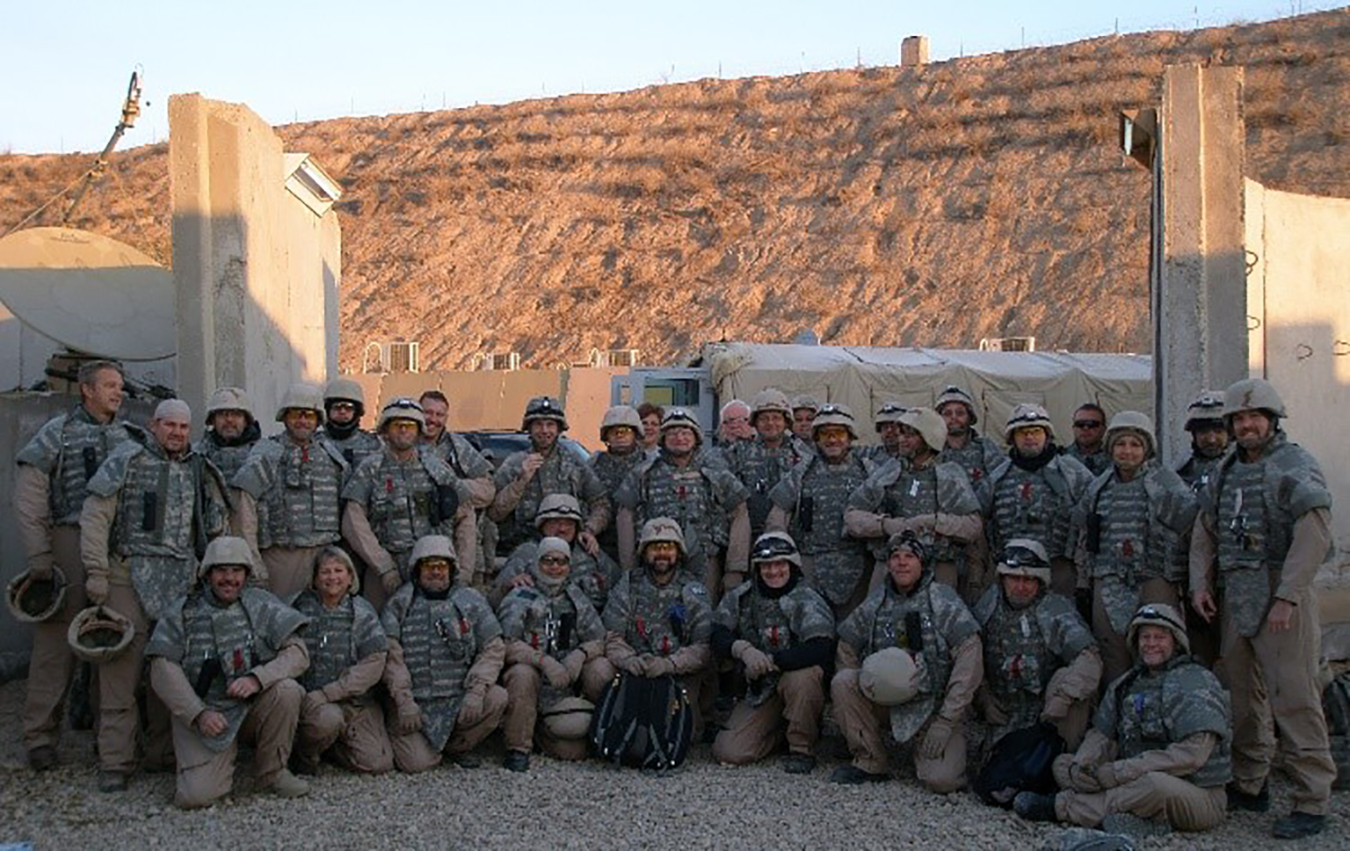 A large group of people in camouflaged uniforms stand outside in front of a masonry wall.