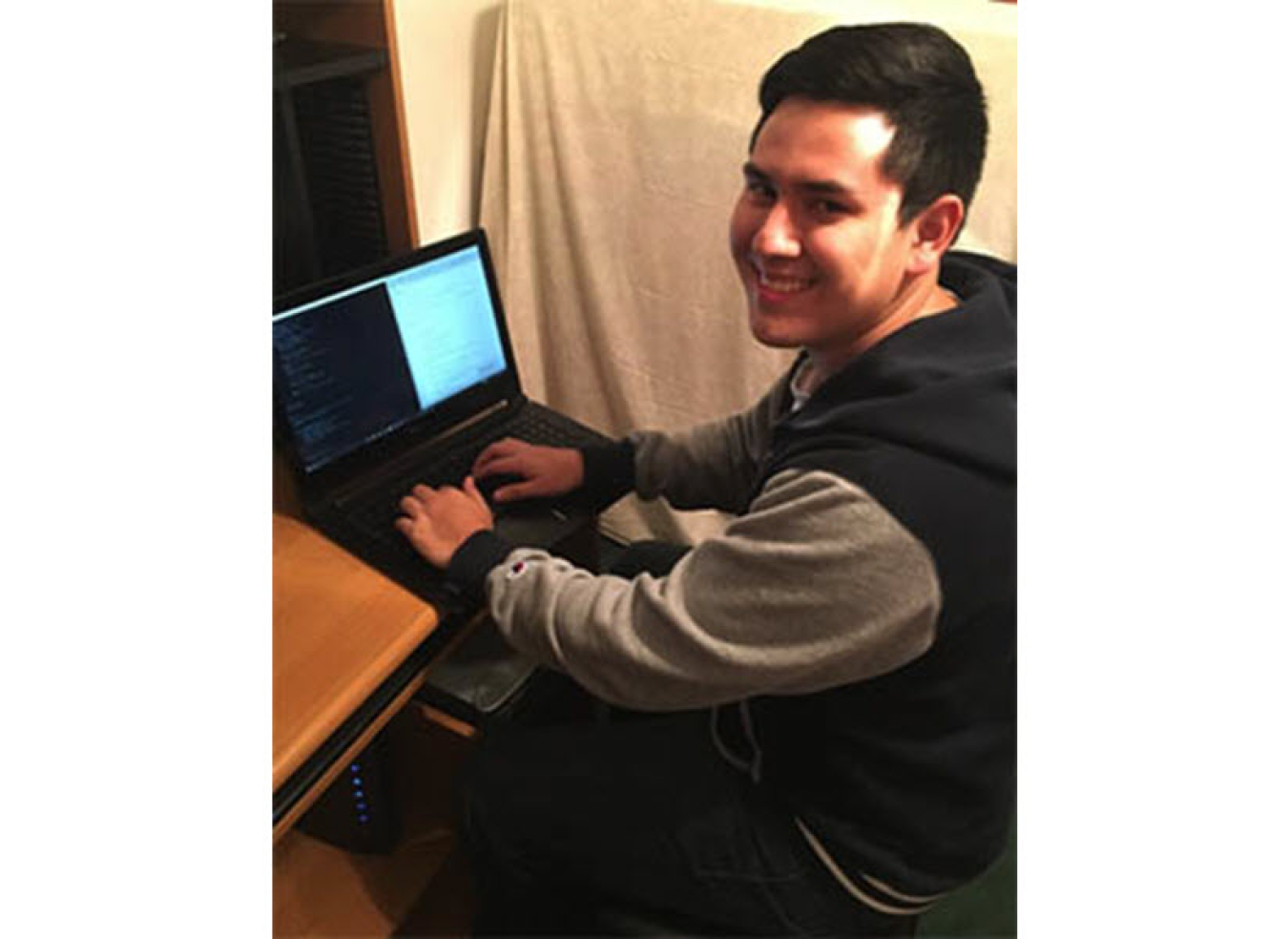 Young man sits at a desk typing on a laptop. He is wearing a navy blue and grey sweatshirt and is smiling with his face turned toward the camera.