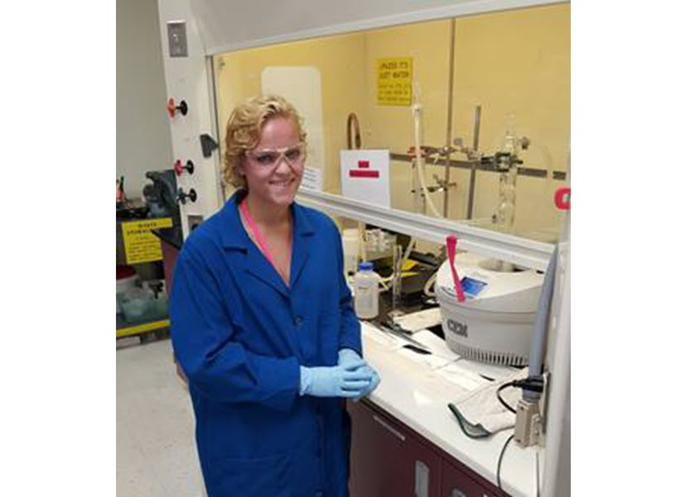 Young woman in a lab smiles at the camera. She is wearing a blue lab coat, blue latex gloves, and safety glasses.