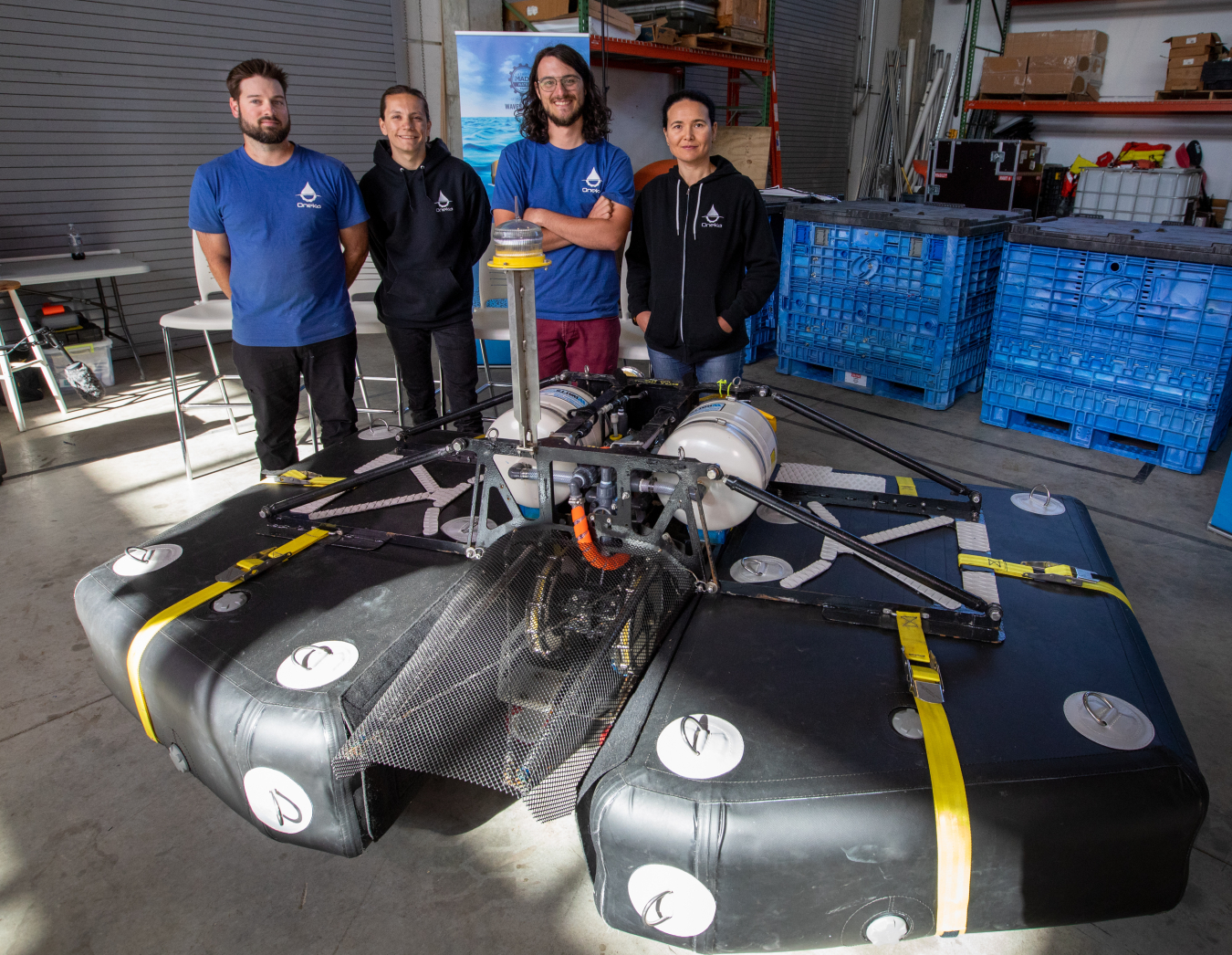 Four members of the Oneka Technologies team standing behind their device, which is as wide as a smart car and has two flotation devices and electronics in between