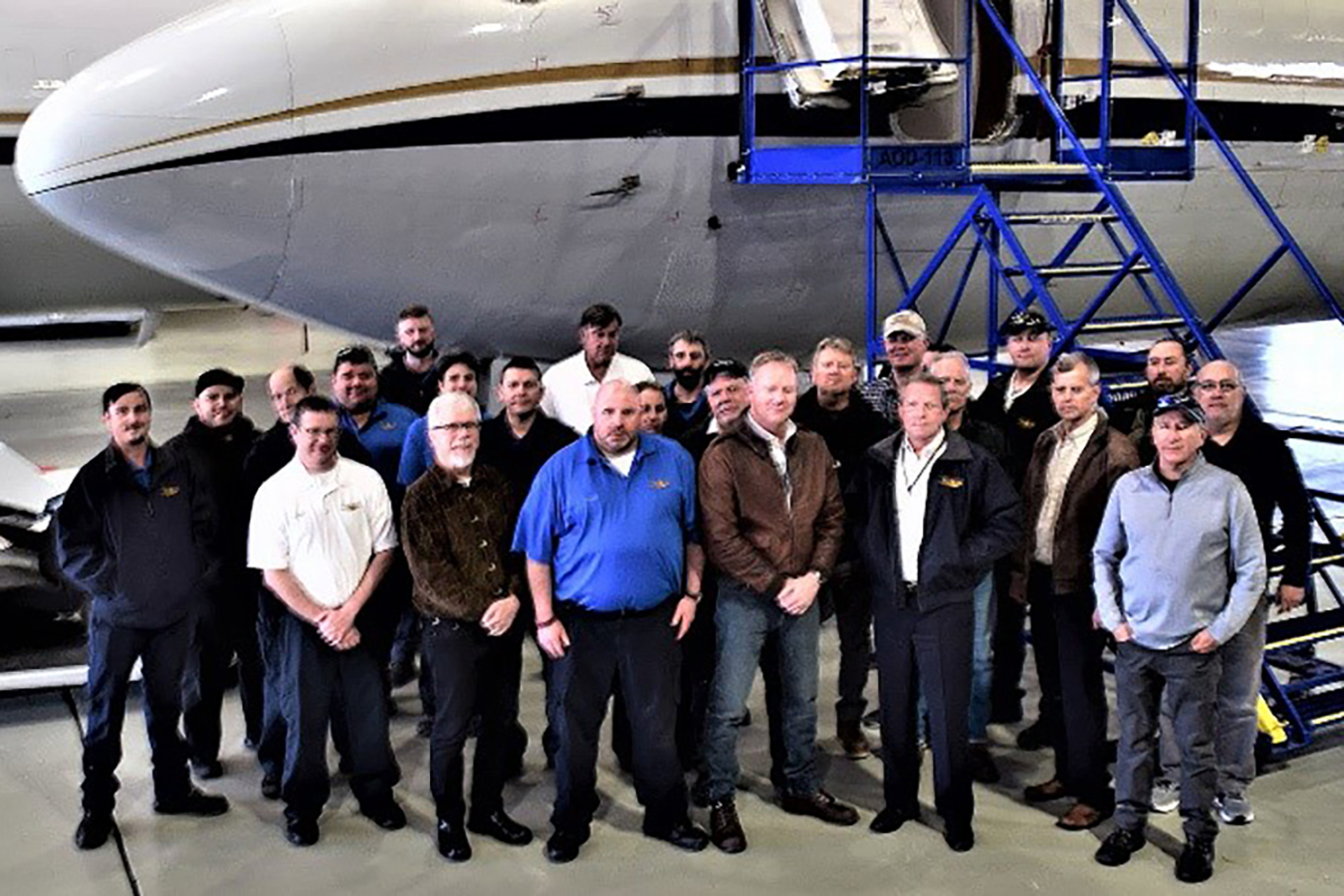 A group of 24 people stand in a hangar in front of an airplane.