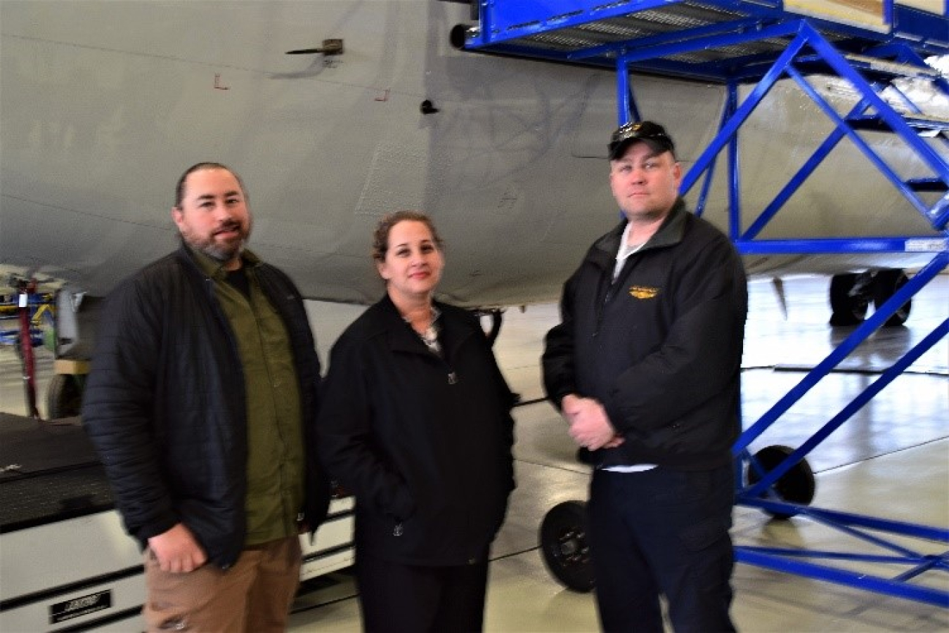 Two men and a woman stand inside a hangar in front of a plane.