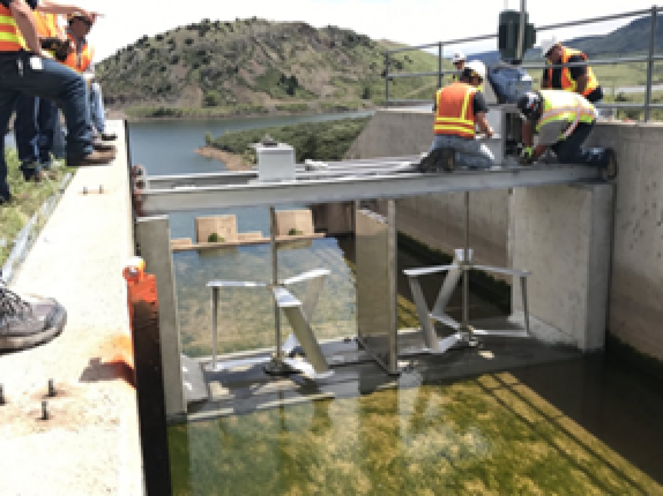 (6)	A photograph of six men wearing hard hats and orange safety vests. The men are standing on two platforms on either side of a small stream. Between the two platforms, a bridge of metal is being constructed. Two men are kneeling on top of this bridge, actively constructing it. 