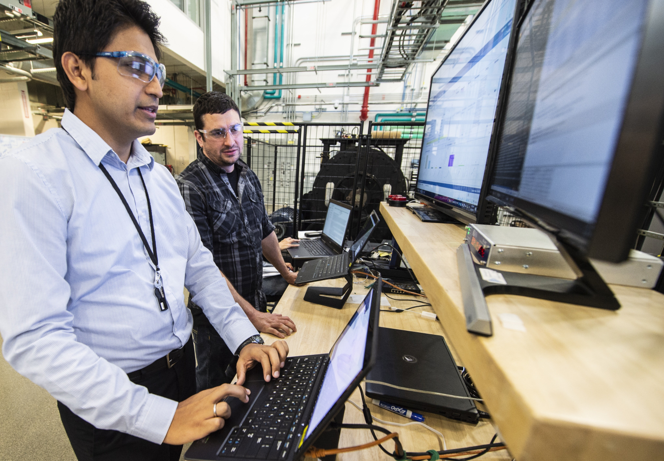 Two researchers standing in front of two large computer screens and two laptops. One of the researchers is moving the mouse on one of the laptops.