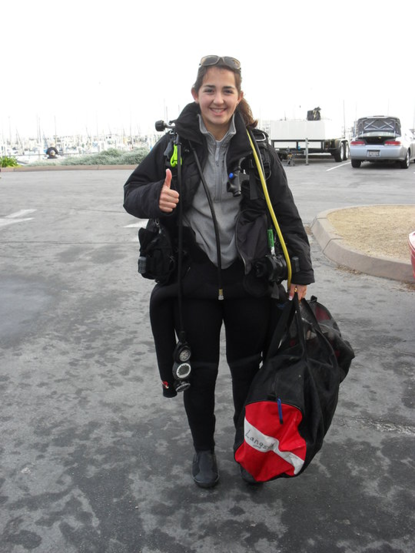 Claire Gonzales poses in a parking lot next to the ocean with her scuba gear giving a thumbs up to the photographer. Claire is wearing some of her gear while other components of the gear are in a bag she is holding. 