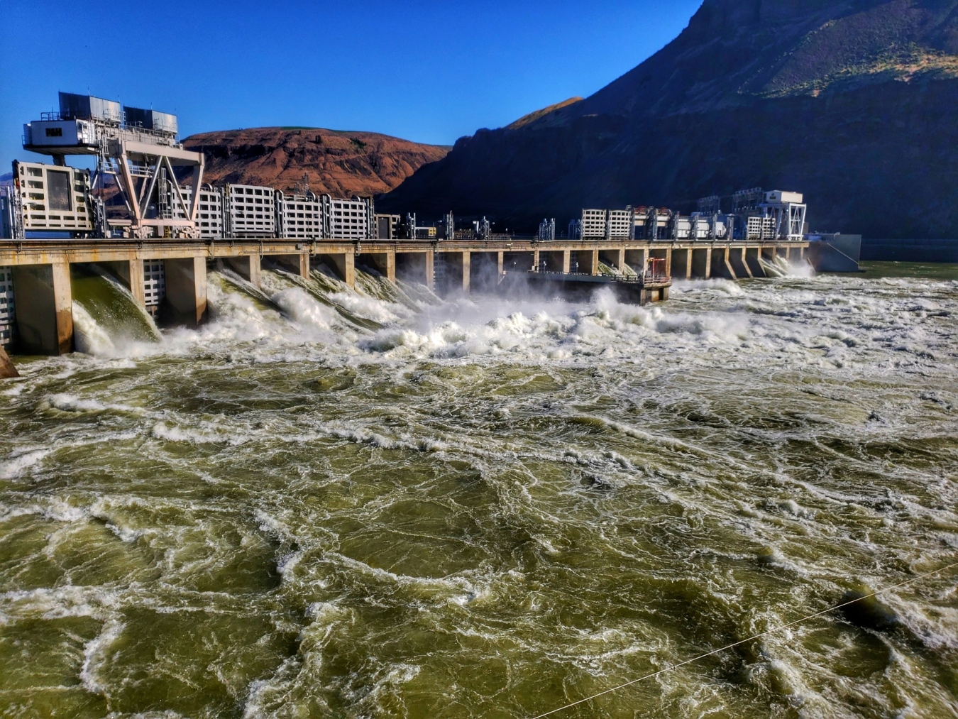 Water rushes through a hydropower facility with reddish hills in the background