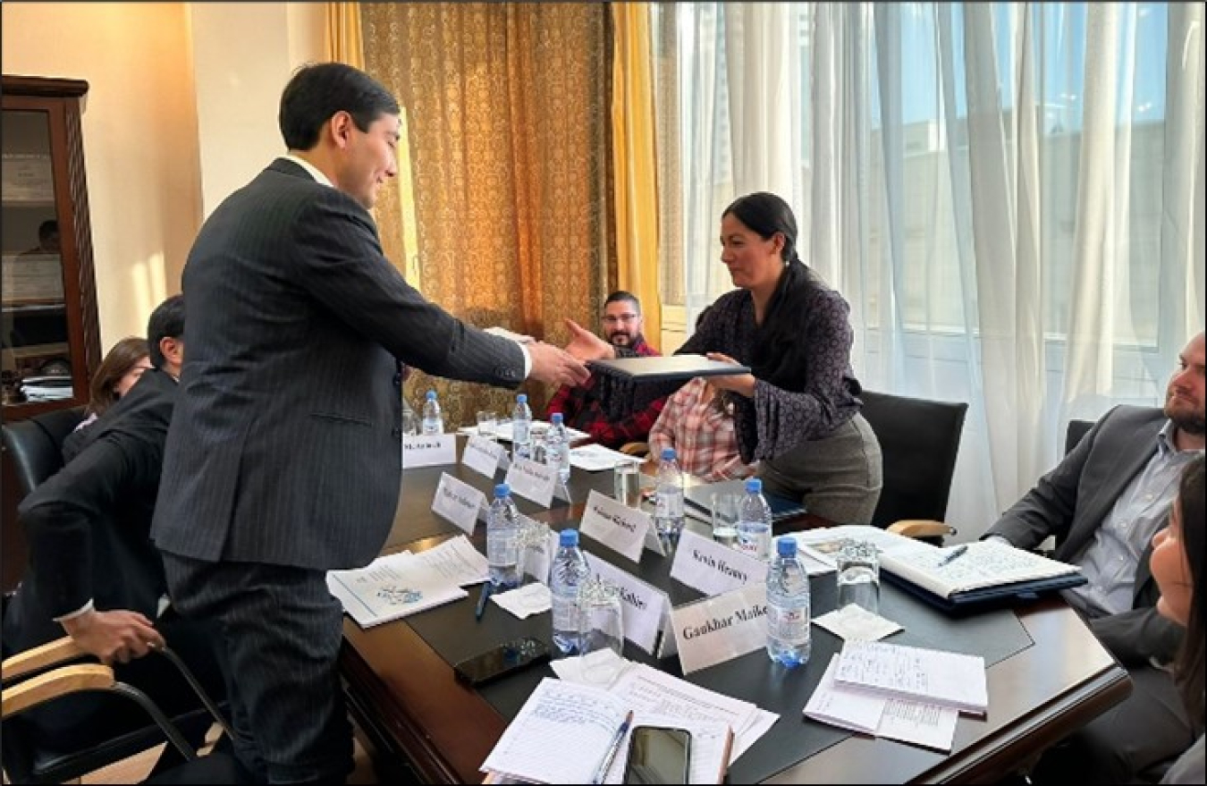 A woman hands a sheaf of papers to a man while they both lean over a conference table. Other people are seated around the table, which has several tentcards with names written on them