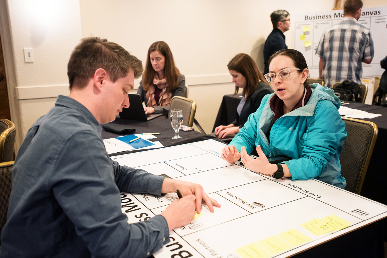 Two people talk while sitting at a table with a poster board on it. 