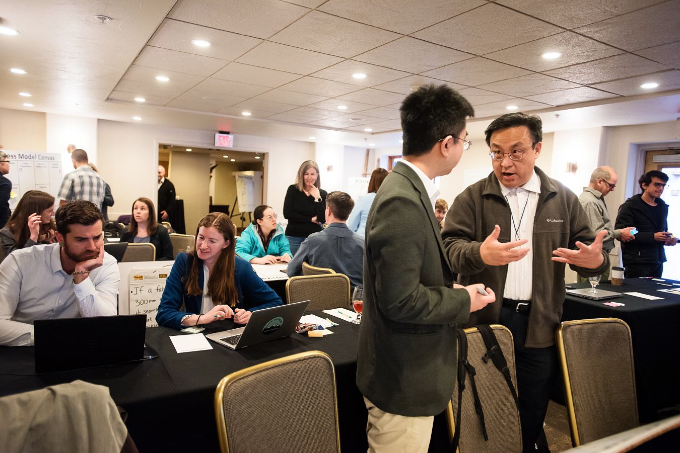 Two men chat in a room of people working at tables.