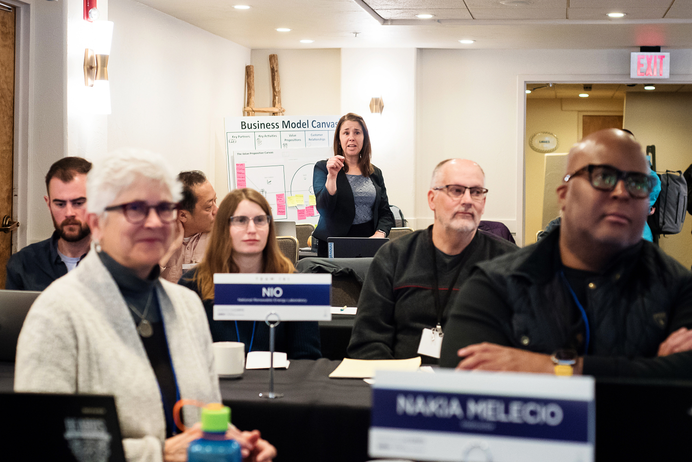 A woman stands to speak from the back of a room with people seated in front of her.