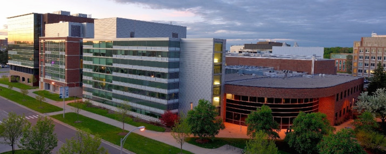 Photo of a tall modern building with large banks of windows and gray stone, with an overcast sky. 