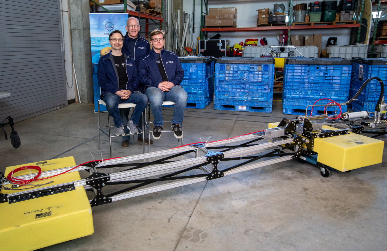 Three people posing behind a yellow and gray wave energy device.