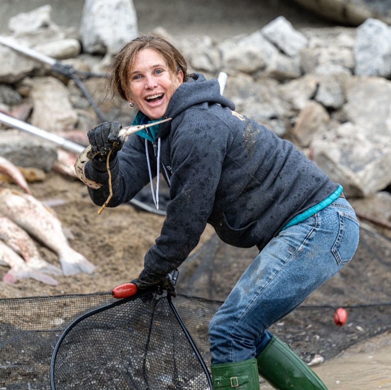 Standing by a hill of dirt and a pile of rocks, Sarah Hill-Nelson holds a net in one hand and a baby longnose gar in the other