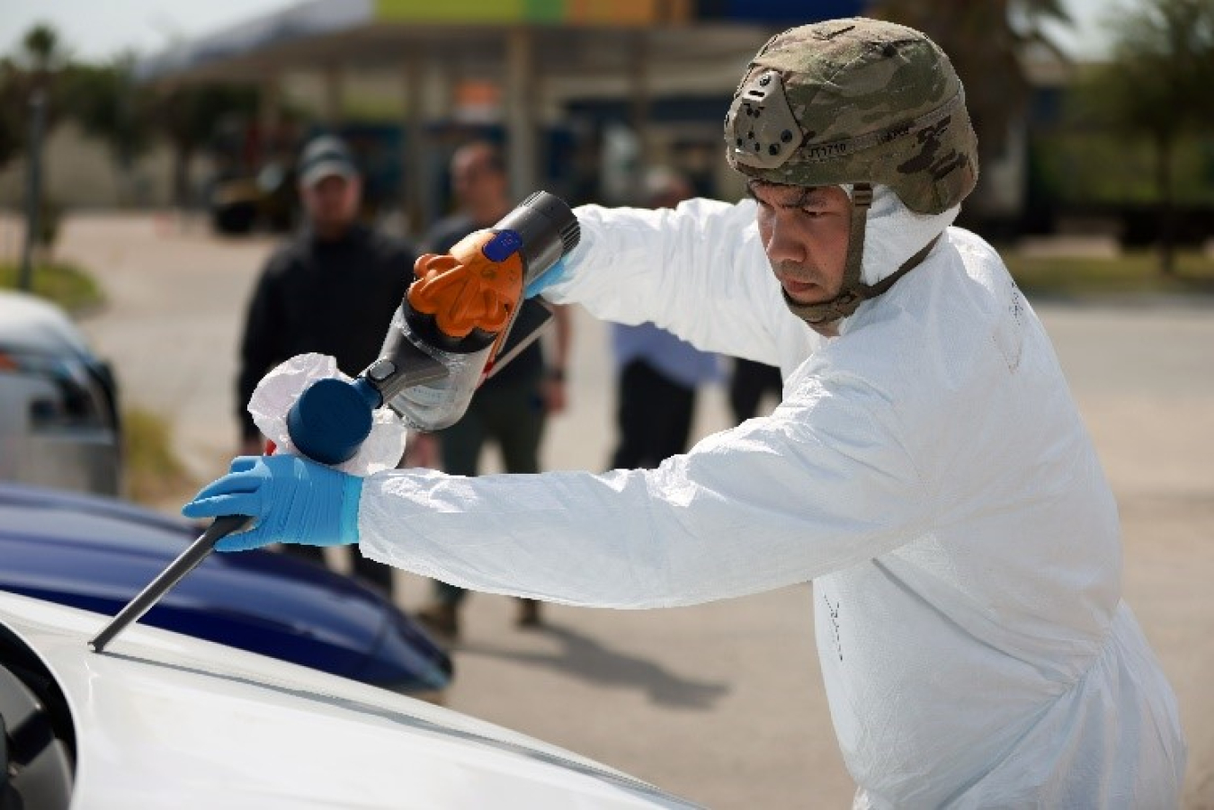 A man in a helmet uses an instrument to collect dust from a car's hood. His face is visible and he is wearing a military-style helmet but the rest of him is covered in radiological protective gear.