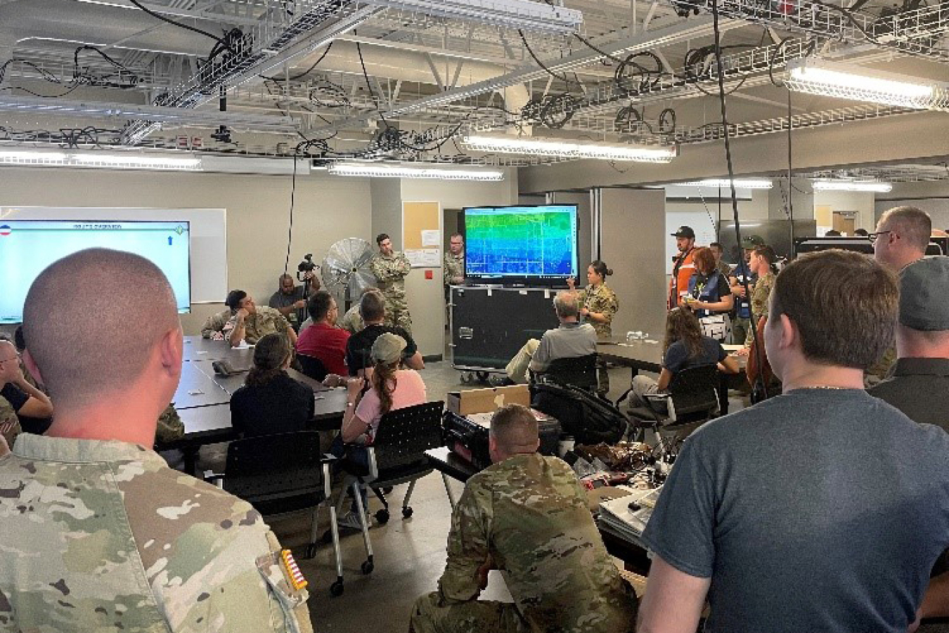 Many people in a room with computer screens, gear, and someone presenting.