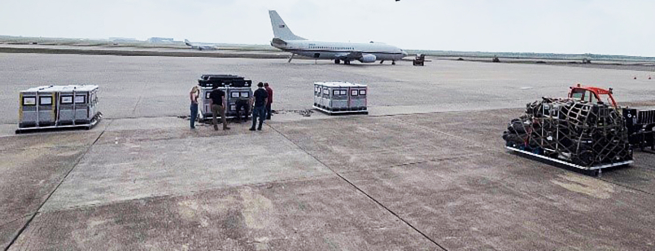 On the tarmac of an airport, three groups of boxes are stacked. A group of four people stand next to one of the stacks. A 737 is parked a hundred yards behind them.