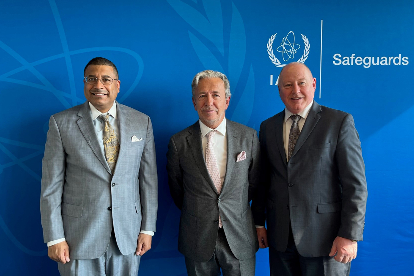 Three men stand in front of a background that has the IAEA Safeguards logo on it.