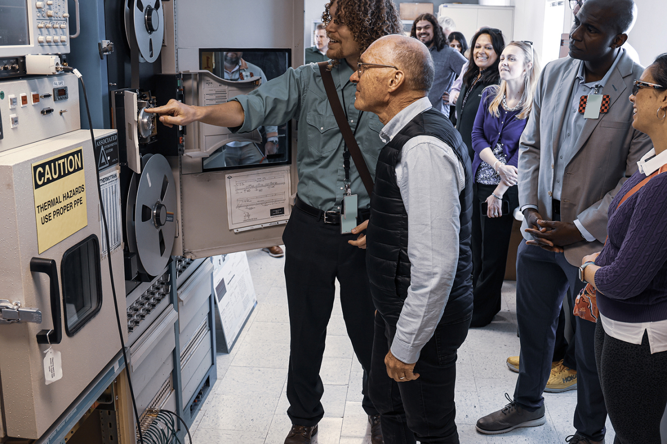 A man talks to Dr. Robert Floyd about a large recording mechanism with prominent reel-to-reel tapes on it.