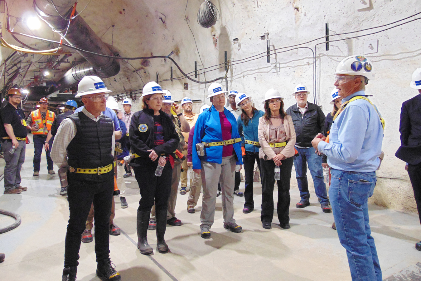 A man explains things to a group of people. It's underground and everyone is wearing hard hats.