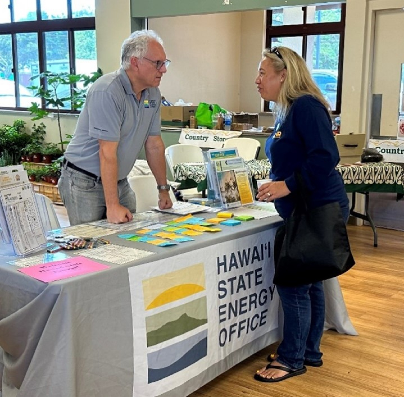 Mark Glick stands behind a table with a sign that reads “Hawai‘i State Energy Office” and talks to a person.