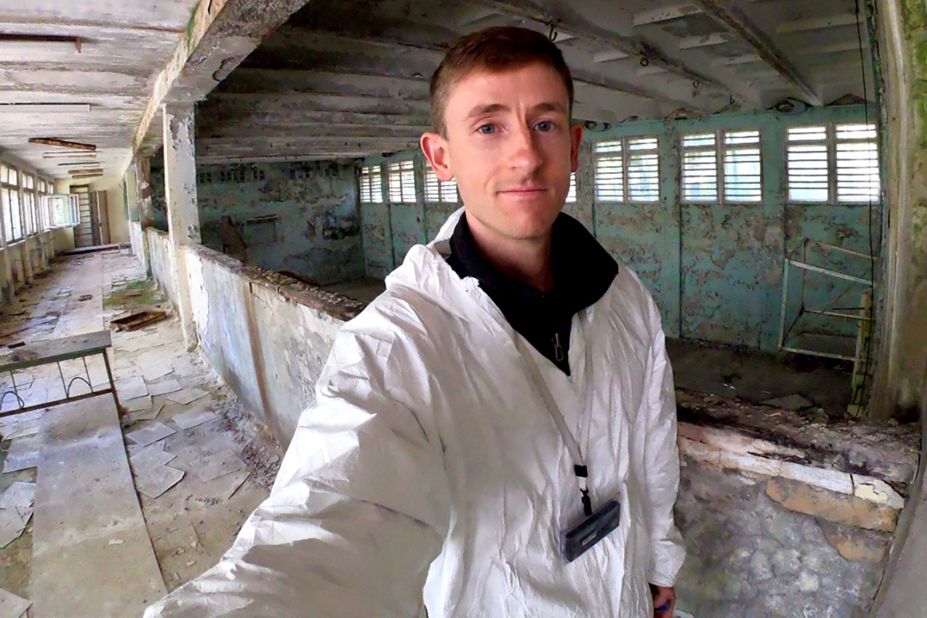 An environmental self-portrait of Adam Stratz. He is dressed in a white protective suit and is standing in a decrepit building.