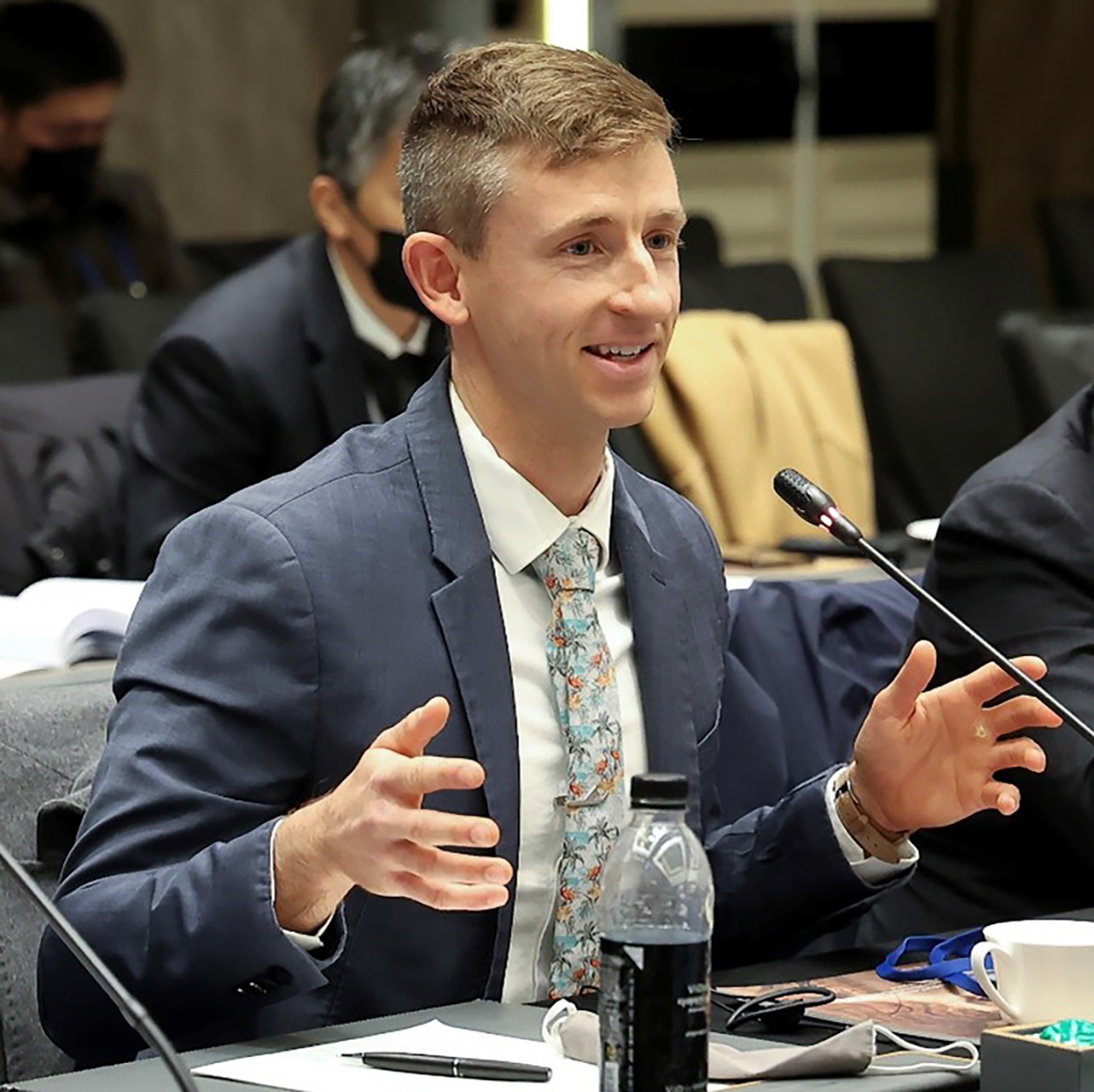 A photo of Adam Stratz while he presents at a desk. He is gesturing with his hands and wearing a suit.