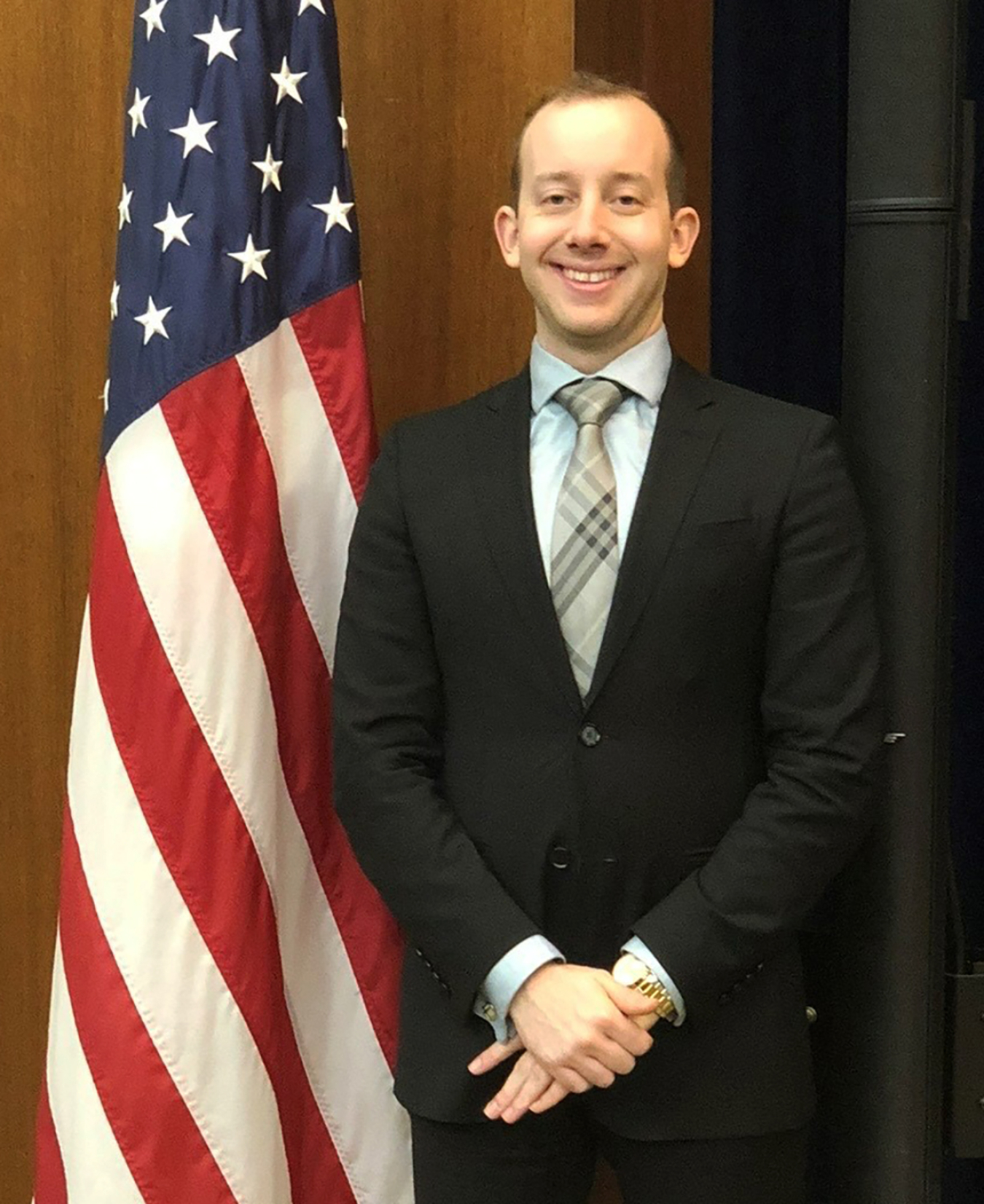 A portrait of Jesse Altum in front of a U.S. flag. He is wearing a dark suit.