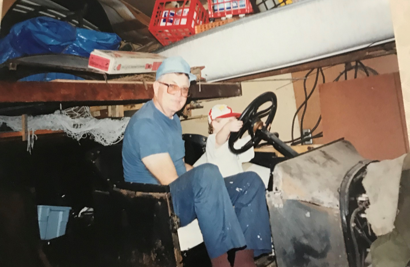 In a garage, a man and a child sit on the front seat of a broken-down early-model car