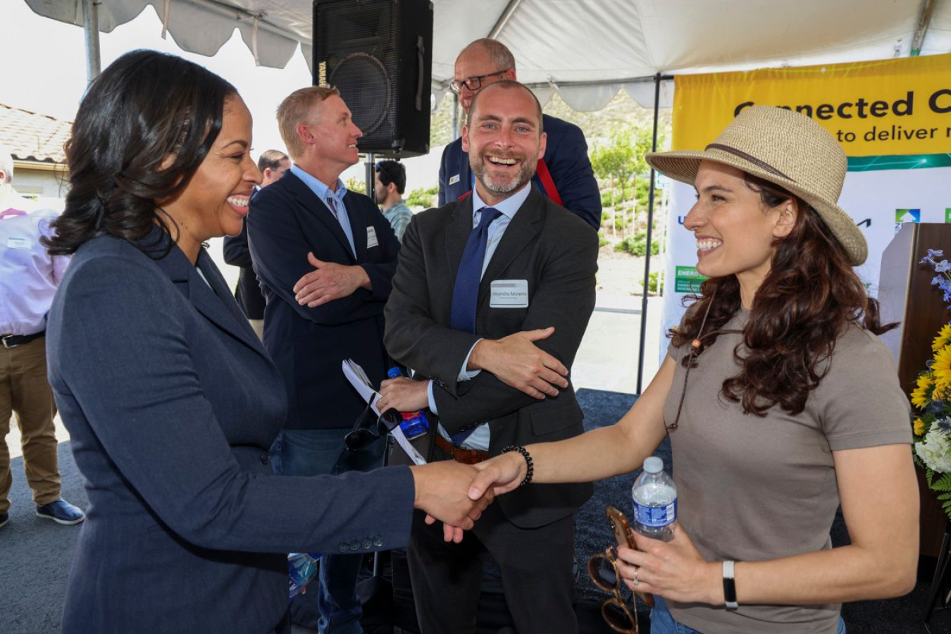 People standing and talking under a tent outdoors: Two women are smiling and shaking hands, while a few men stand nearby.