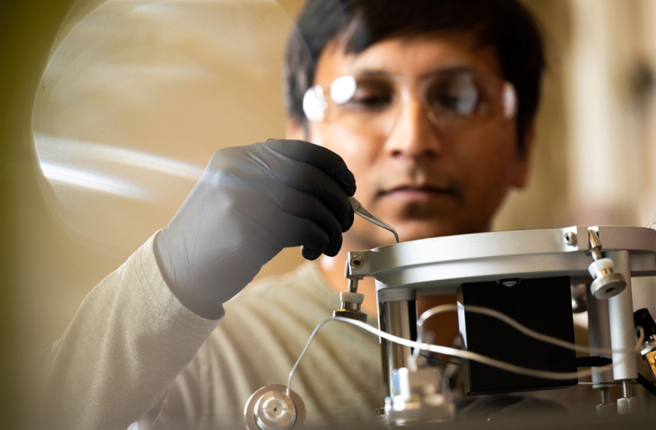 Vimal Balasubramanian (an East Asian man in lab gloves) using a curved metal tool on an isotope ratio mass spectrometer at the EMSL user facility.