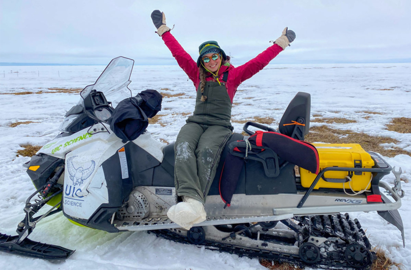 Serina Wesen (a white woman in snow overalls, snow boots, and snow gloves) sitting on a snow machine full of gear on the tundra with melting snow