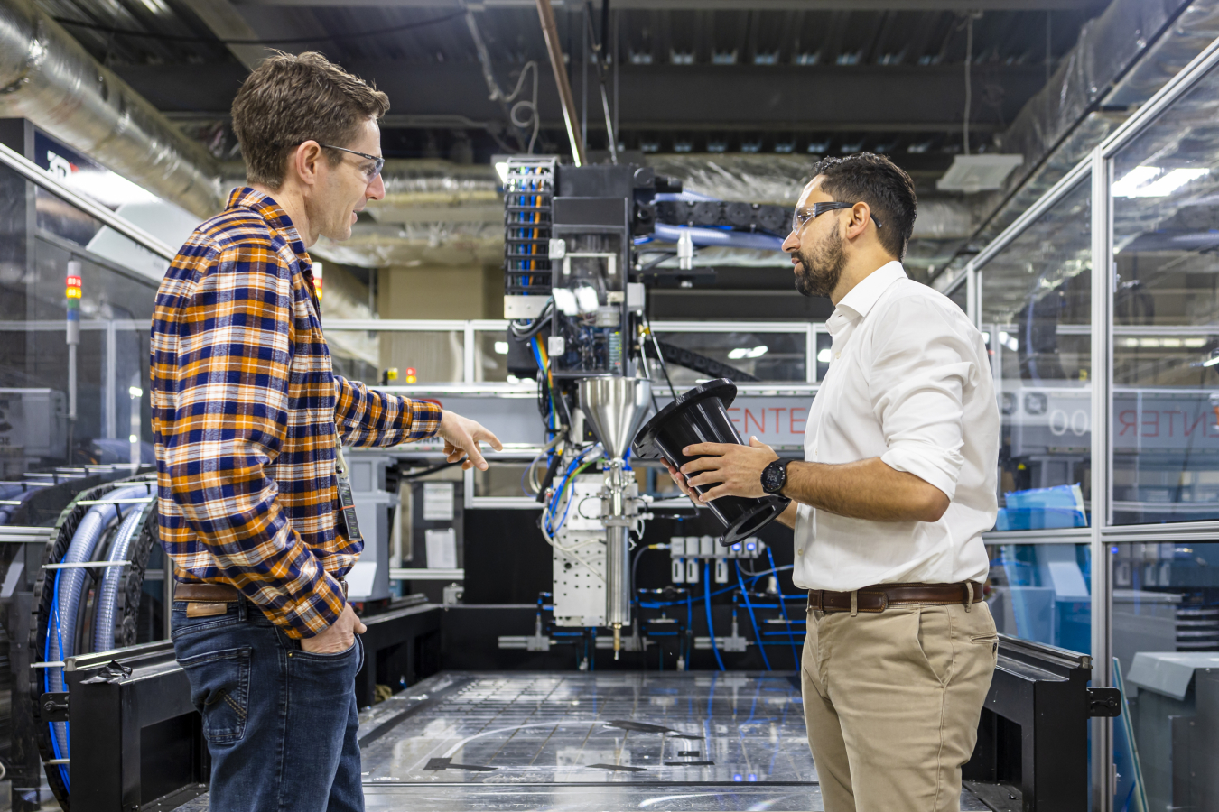 Mirko Musa and Jesse Heineman chat while wearing goggles and standing in a warehouse with mechanical devices] 