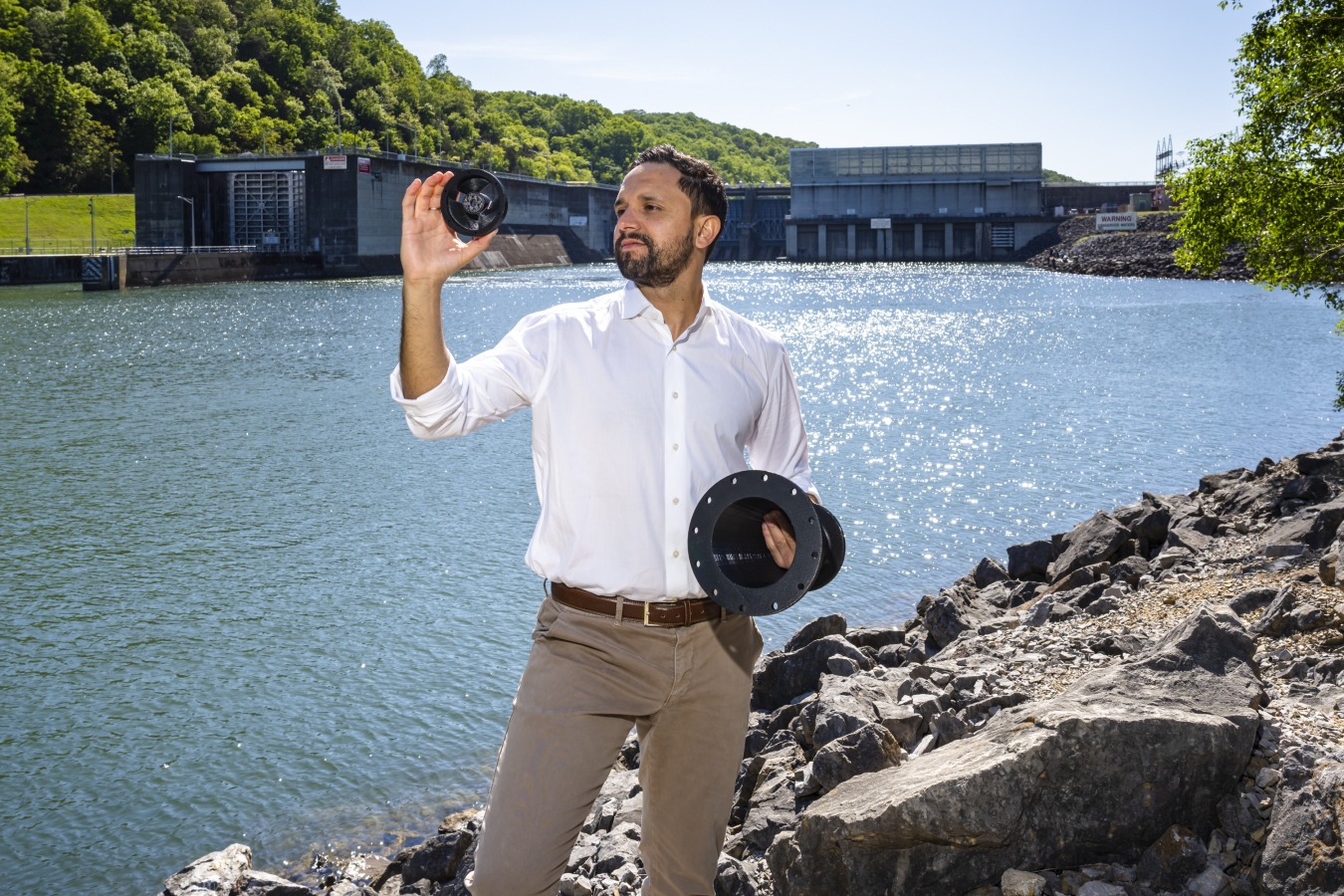 Mirko Musa holds up 3D printed hydropower turbine models while standing on a rocky shoreline of a river with a dam in the background