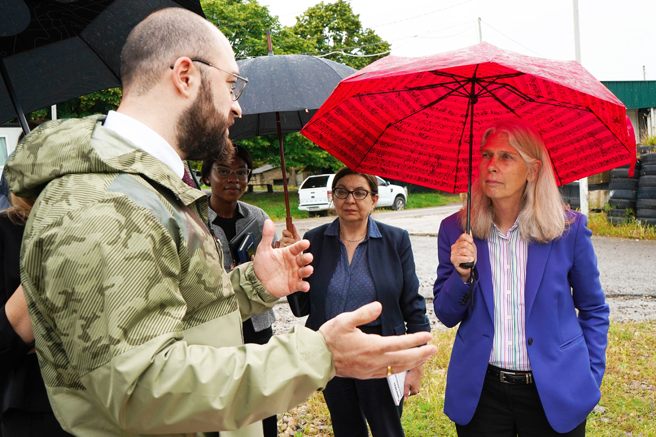 NNSA Administrator Hruby, holding a red umbrella, listens to a man wearing military fatigues.