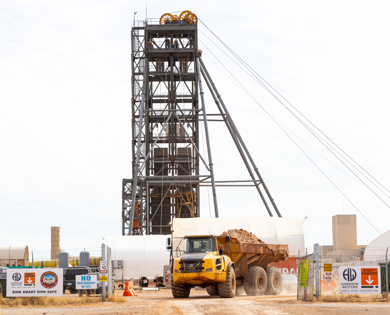 A truck hauls excavated salt away from the Waste Isolation Pilot Plant's (WIPP) utility shaft project, marked by a large aboveground steel headframe. The shaft, key to WIPP’s new air ventilation project, has reached the depth necessary to allow horizontal tunneling work to begin on connecting the shaft to the WIPP underground repository complex. The repository is 2,150 feet below ground level.