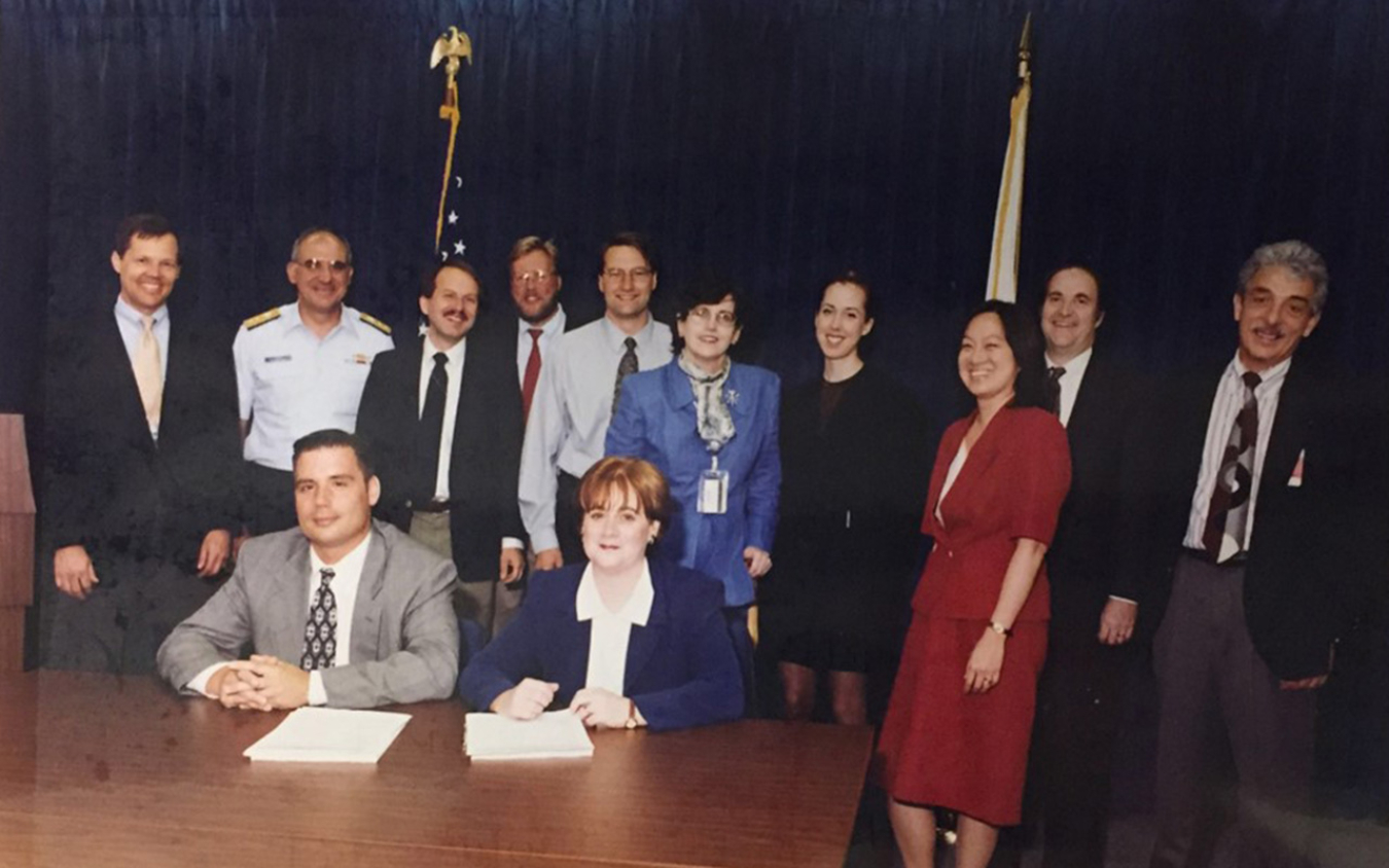 Two people sitting at a table with two stacks of paper next to them. A group of individuals stand behind them along with the U.S. and Alaska state flags.